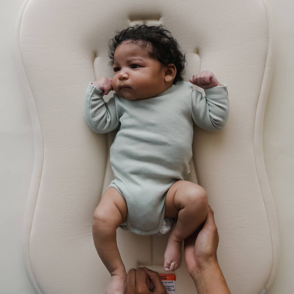 Baby in a light green onesie lying on a white cushion, held by an adult's hands.