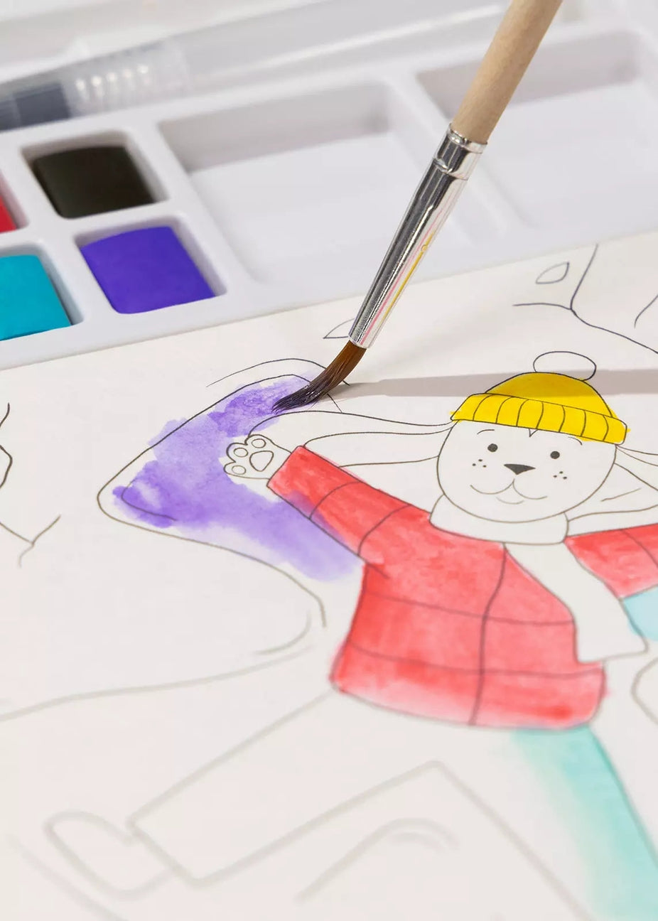 A close-up of a child's watercolor painting in progress, showing a cartoon bear character being painted with a paintbrush. The bear is wearing a yellow beanie and a red jacket, with purple and teal colors being applied.