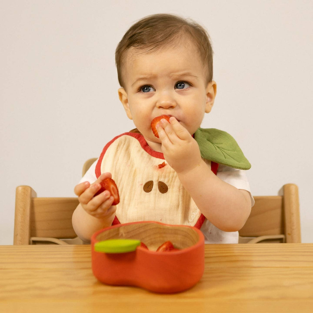 Baby eating a tomato with a bib on, sitting at a table.