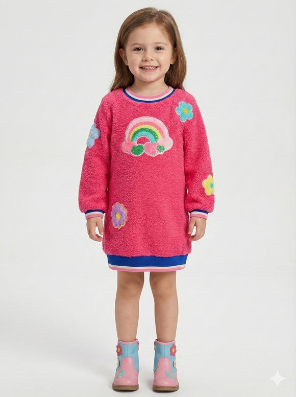 A young girl stands against a light gray studio background wearing a bright pink fuzzy long-sleeve dress with a rainbow and flower applique design, blue and pink trim, and pink boots.