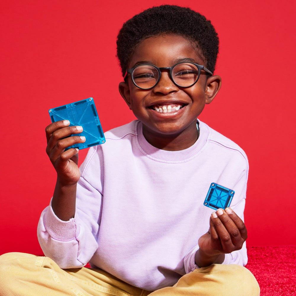 A smiling child wearing glasses and a light purple sweatshirt holds up small translucent blue magnetic tiles in each hand against a vibrant red background.