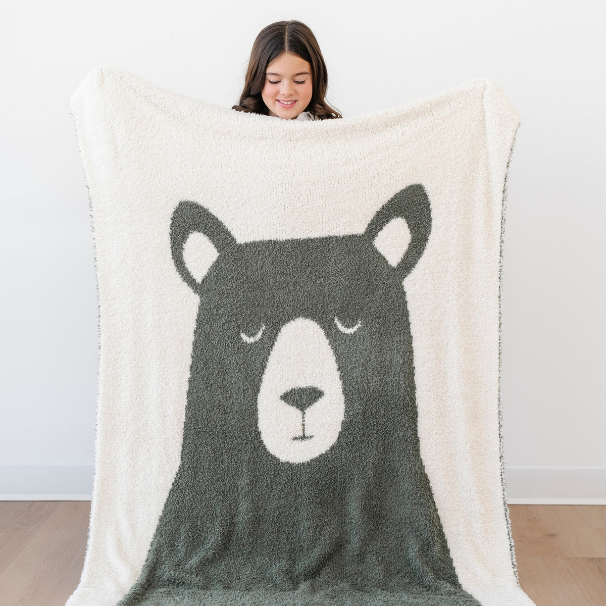 A person holds up a large cream sherpa blanket featuring a dark gray bear face design, with the person's head visible above the blanket against a plain white wall.