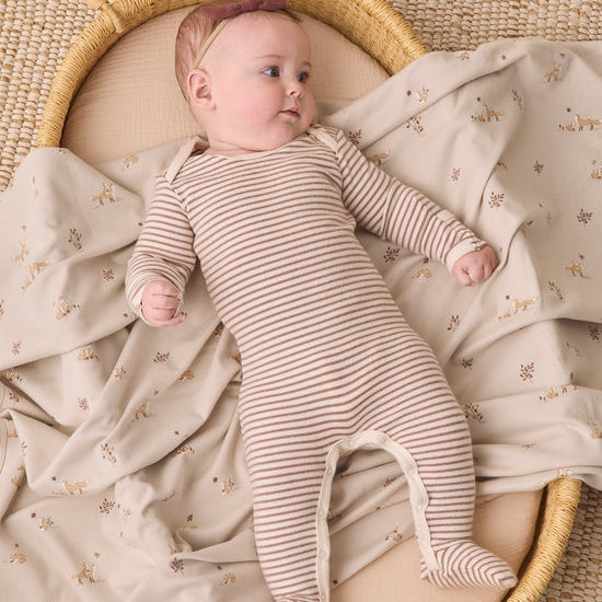 A baby lying on a cream and beige floral blanket inside a woven rattan bassinet, wearing a beige and brown striped footed sleeper.