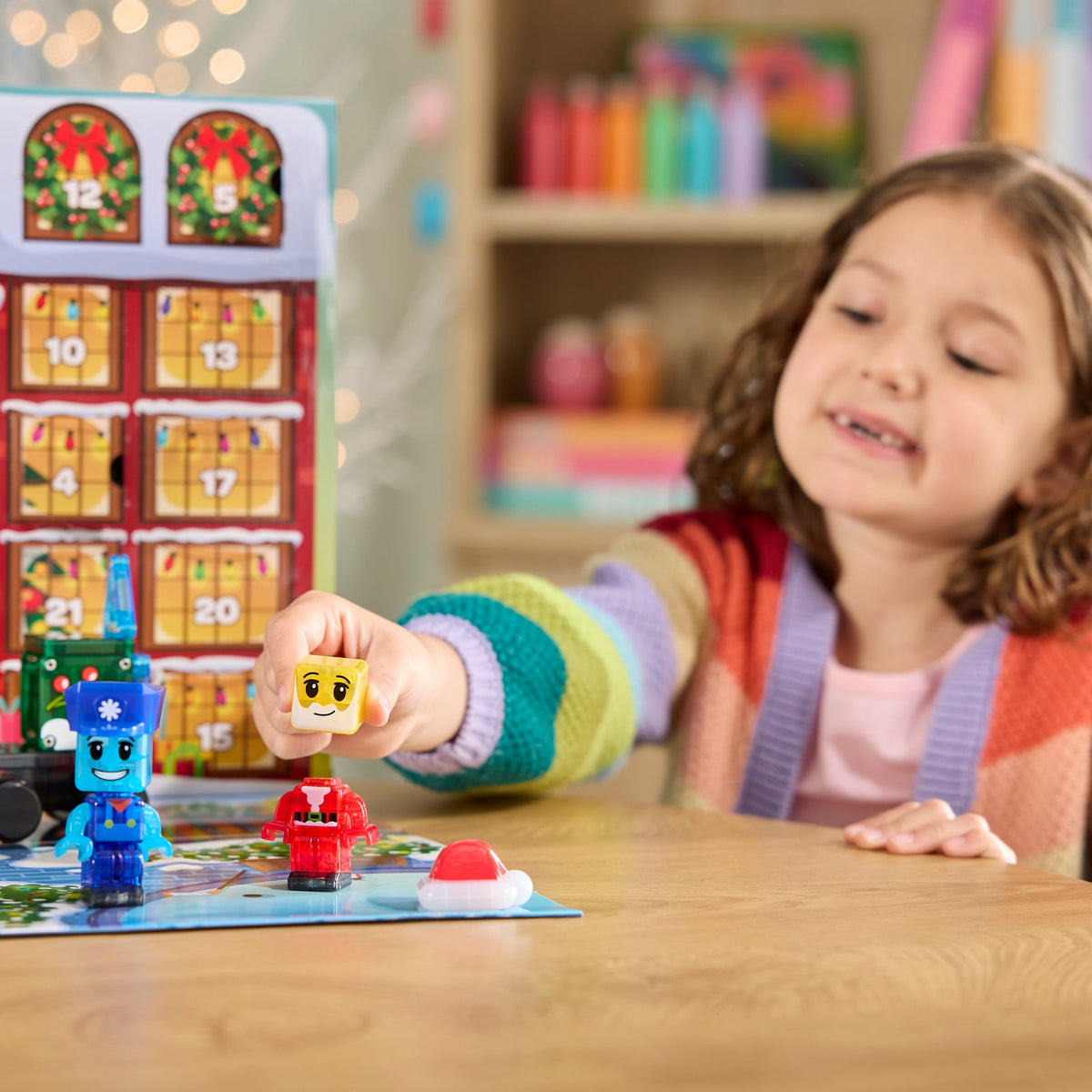A young girl with curly brown hair smiles while playing with a toy Advent calendar and small character figures on a wooden table. The colorful calendar stands behind her, and the room background is softly blurred with shelves and lights.