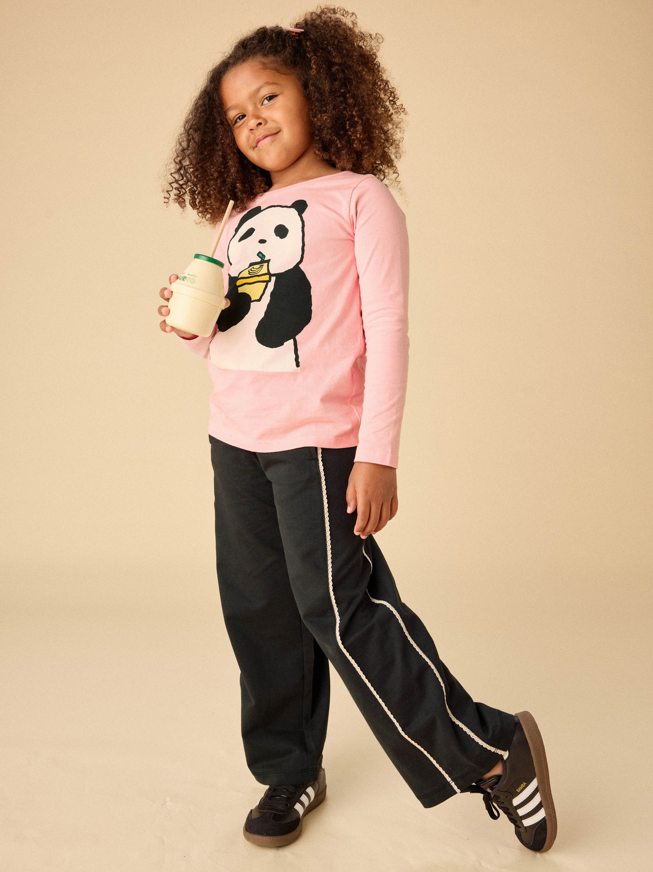 Child wearing a pink shirt with a panda design and black pants, holding a drink.