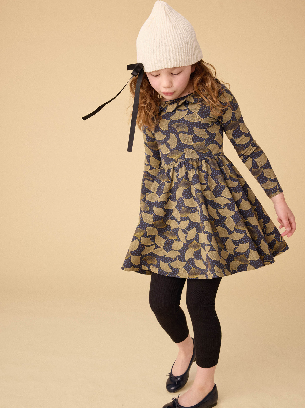 Young girl wearing a patterned dress with long sleeves and a white knit hat on a beige background