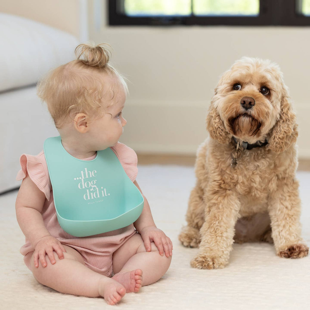 Baby wearing a bib with text sitting next to a dog on a light-colored floor.