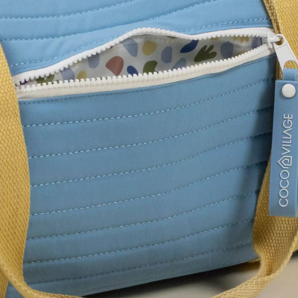 Close-up of a light blue quilted bag with a white zipper, a multicolor patterned interior lining, and a yellow fabric strap. A blue hangtag on the right side reads COCO VILLAGE.