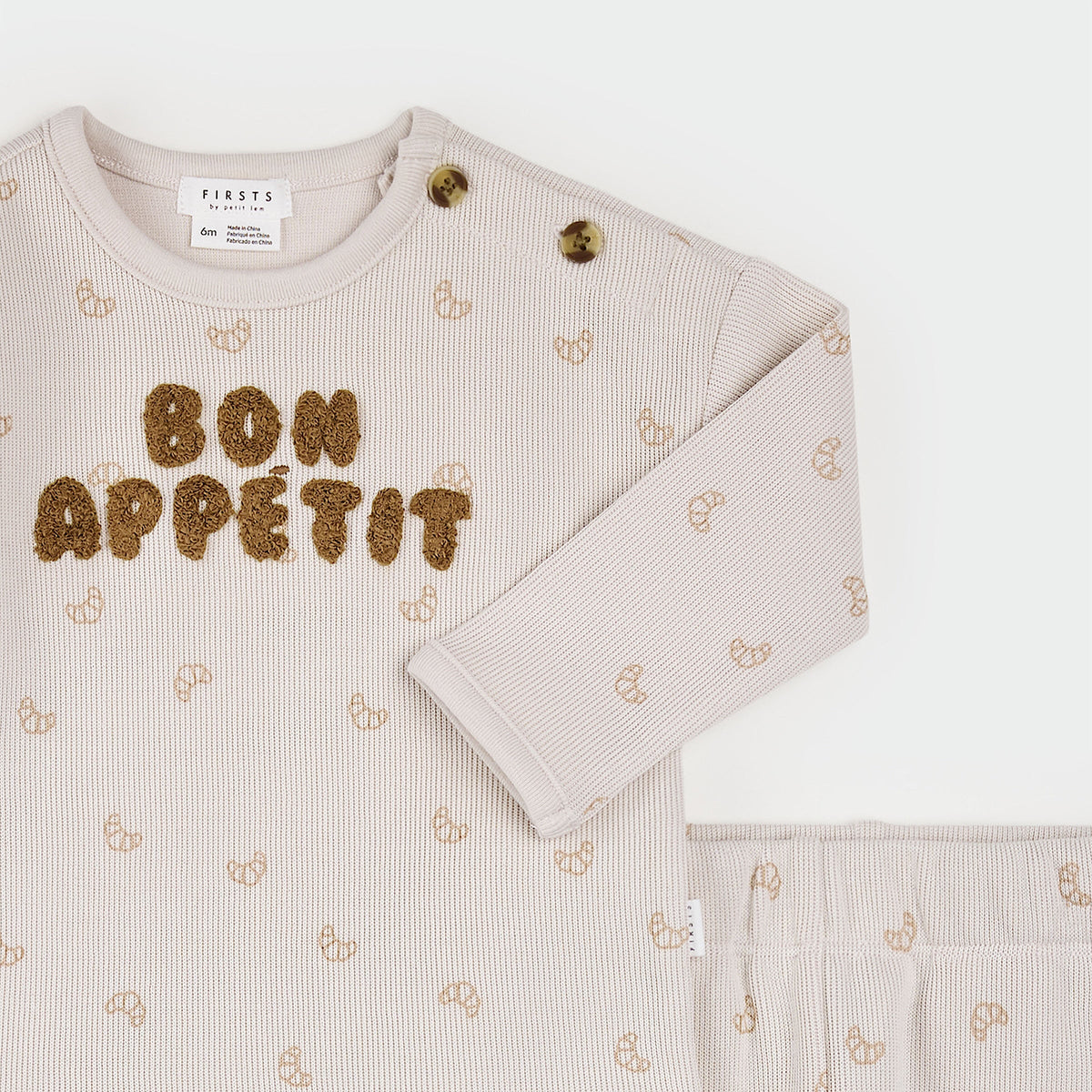 A baby long-sleeve top and matching pants set in a soft, light beige ribbed fabric, featuring a 'BON APPÉTIT' text design in brown sequins on the chest and a subtle repeating bow pattern. The brand 'Finsts' is visible on the inner neck label.