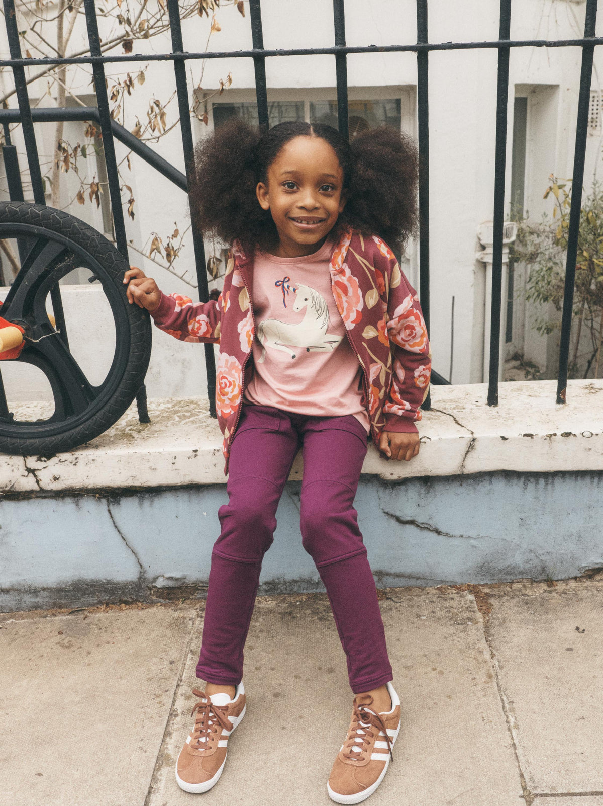 A young girl with dark curly hair sits on a low wall outdoors, wearing a pink graphic t-shirt under a floral patterned jacket, magenta pants, and brown sneakers.