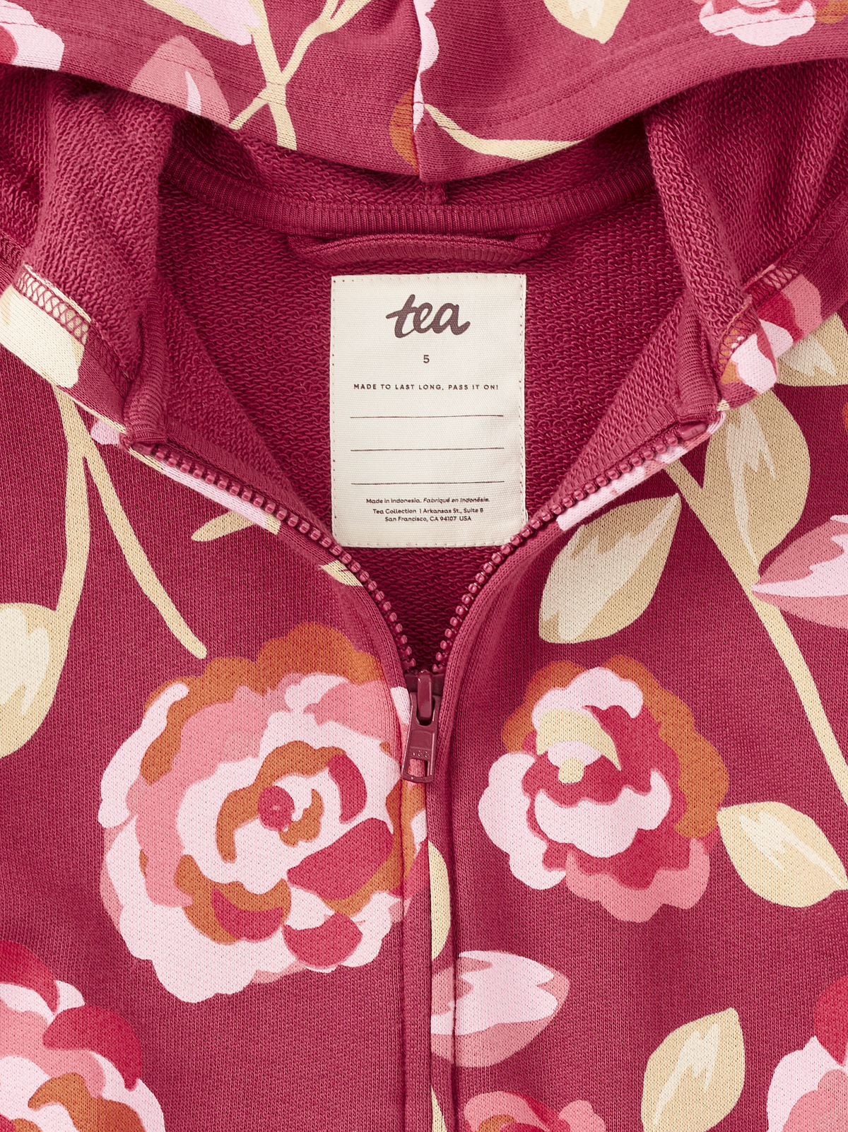 Close-up of a zip-front hooded sweatshirt with a floral print in dusty rose, pink, cream, and muted orange. A white neck label reads tea.