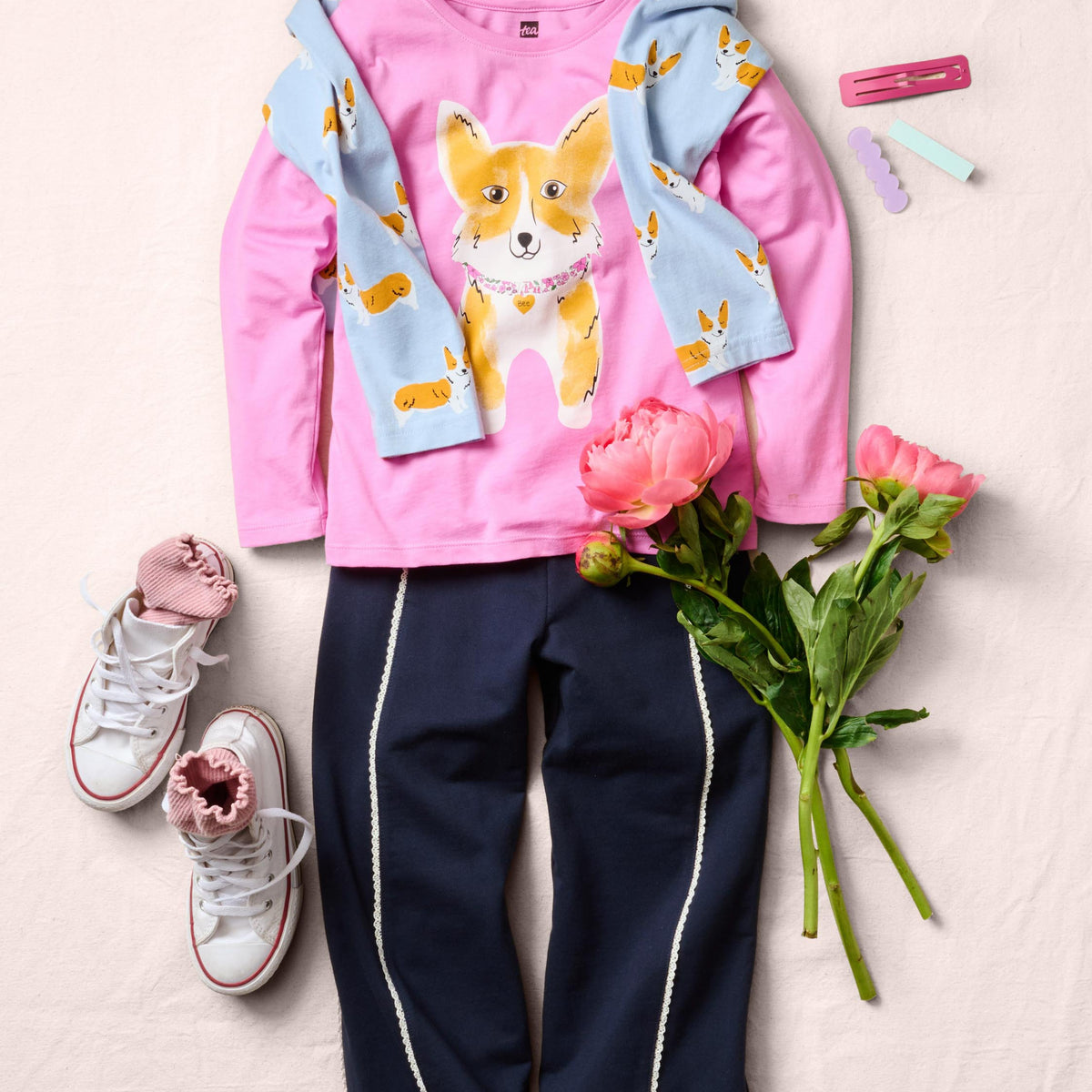 A children's outfit laid flat on a light beige fabric background, featuring a pink long-sleeve top with a corgi graphic and light blue patterned sleeves, navy pants with white side stripes, pink sneakers, and pink flowers as styling props.