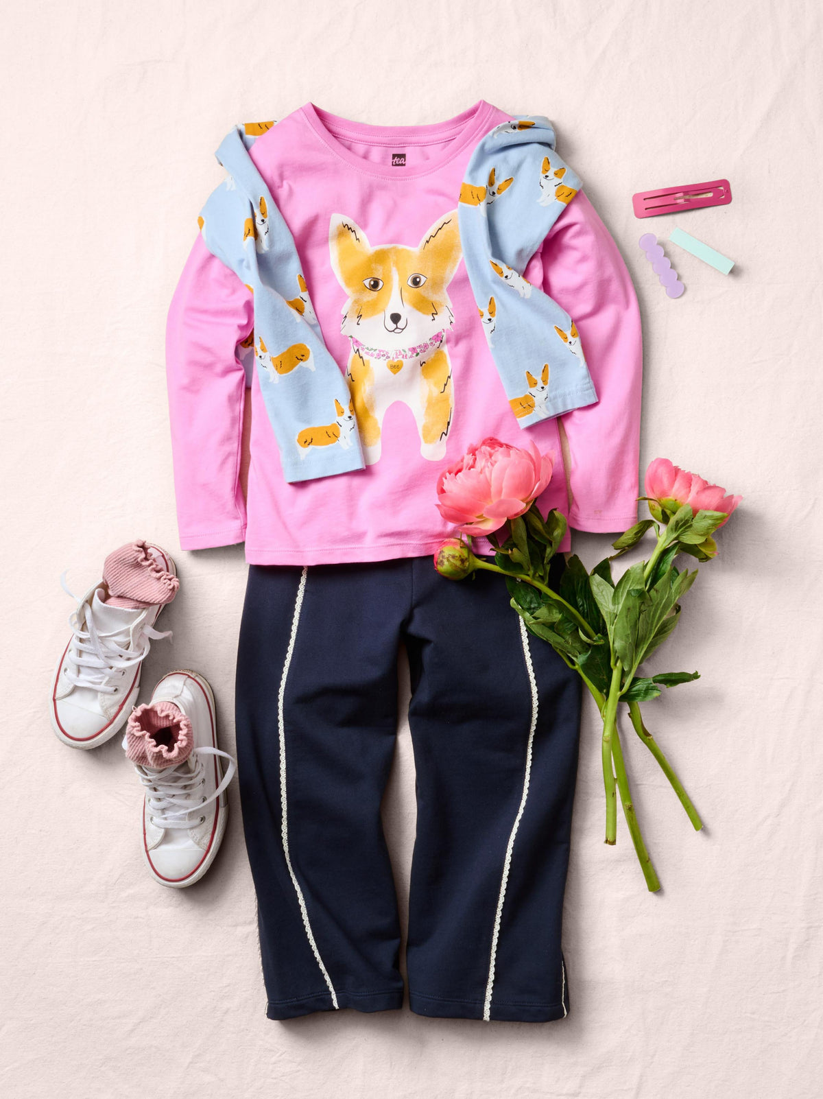 A children's outfit laid flat on a light beige fabric background, featuring a pink long-sleeve top with a corgi graphic and light blue patterned sleeves, navy pants with white side stripes, pink sneakers, and pink flowers as styling props.