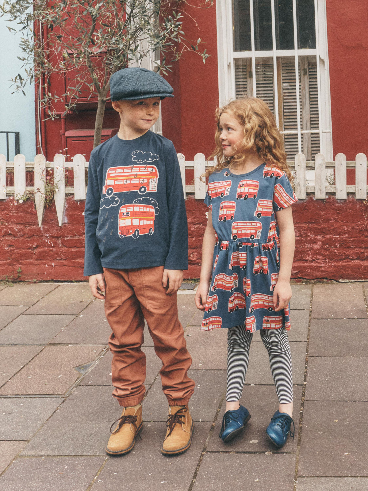 A boy and a girl standing on a paved street in front of a red brick building with a white picket fence. The boy wears a navy blue long-sleeve shirt with a red double-decker bus print, brown pants, and a navy blue flat cap.
