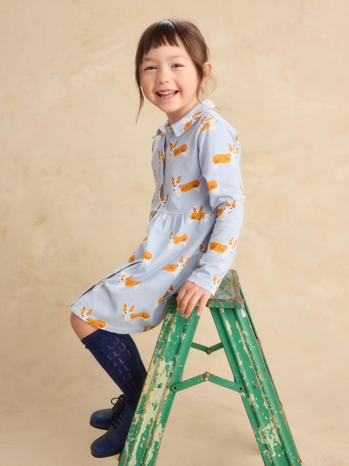 A young girl with brown hair sits on a distressed green wooden step ladder, wearing a light blue long-sleeve dress with an orange bunny print, dark blue knee-high socks, and navy boots against a textured beige studio backdrop.