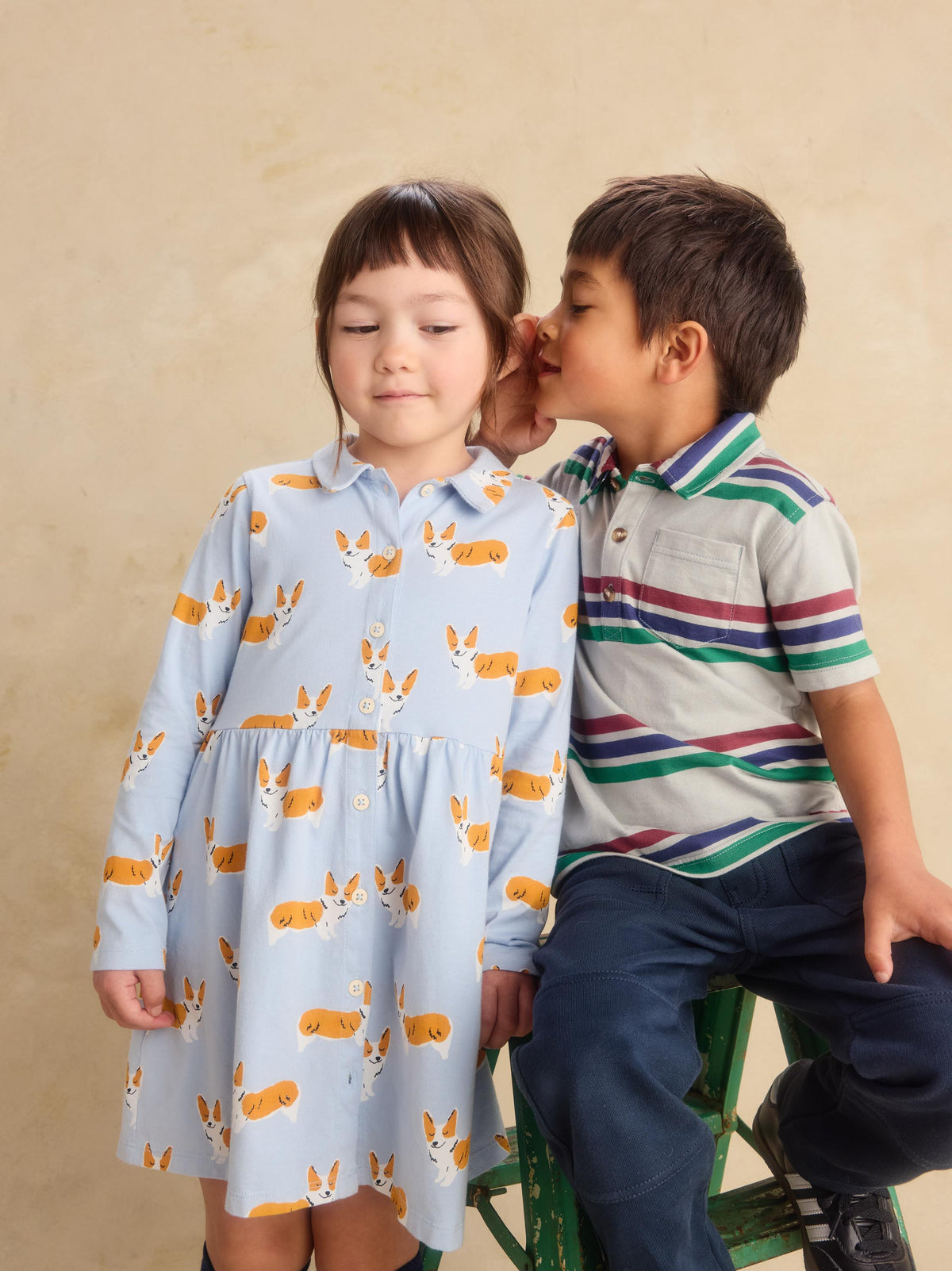 A young girl in a light blue collared dress with a corgi print sits next to a boy in a striped polo shirt who is whispering in her ear, set against a warm beige background.