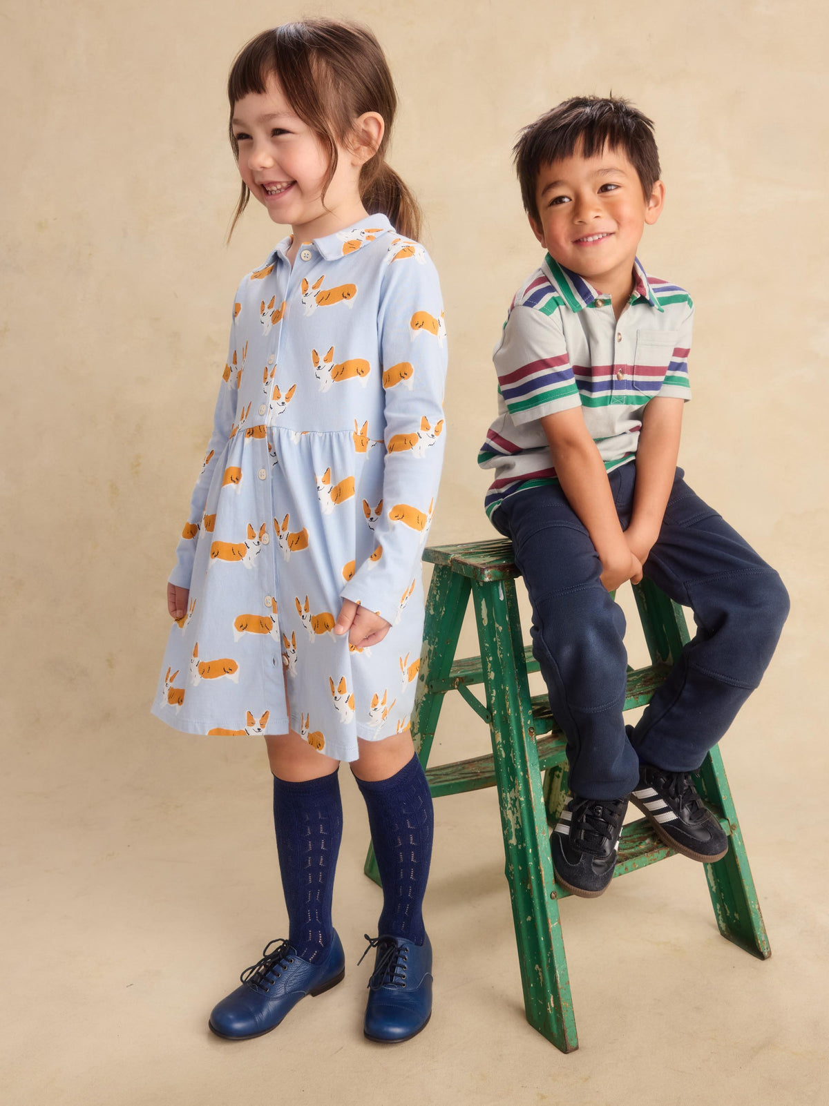 A young girl stands wearing a light blue long-sleeve dress with a yellow and white animal print, paired with dark blue knee-high socks and blue shoes. A young boy sits on a green wooden step stool wearing a striped polo shirt, navy pants, and black sneakers.
