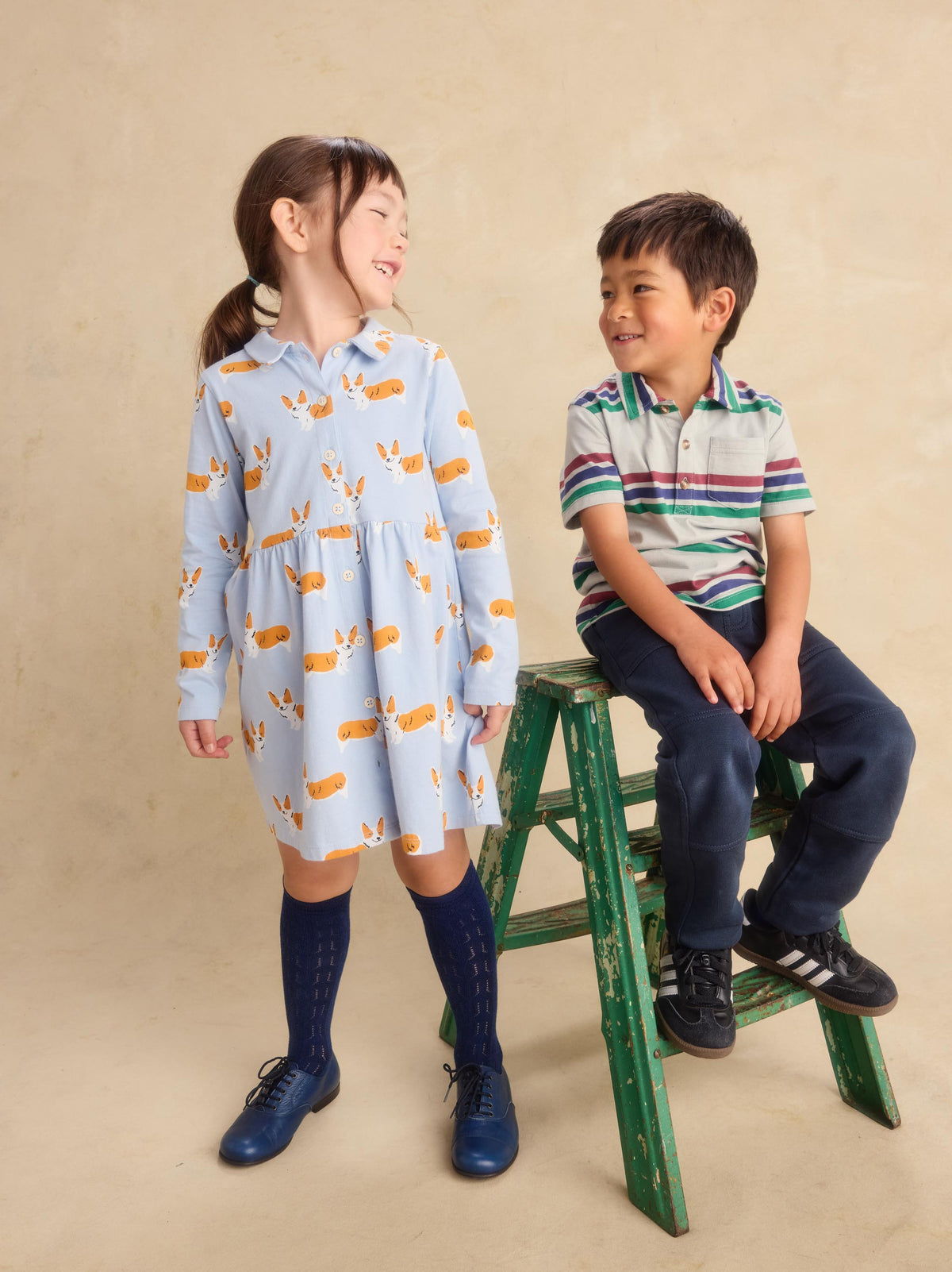 A young girl stands wearing a light blue collared dress with an orange and yellow animal print, paired with dark blue knee-high socks and blue shoes. A young boy sits on a green wooden step stool wearing a green, white, and blue striped polo shirt and navy pants.