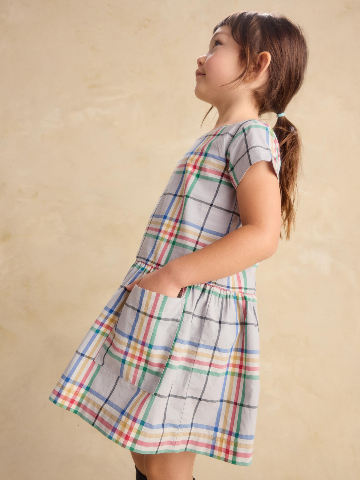 A young girl with brown hair in a ponytail is wearing a short-sleeved plaid dress with pockets, standing against a textured beige background.