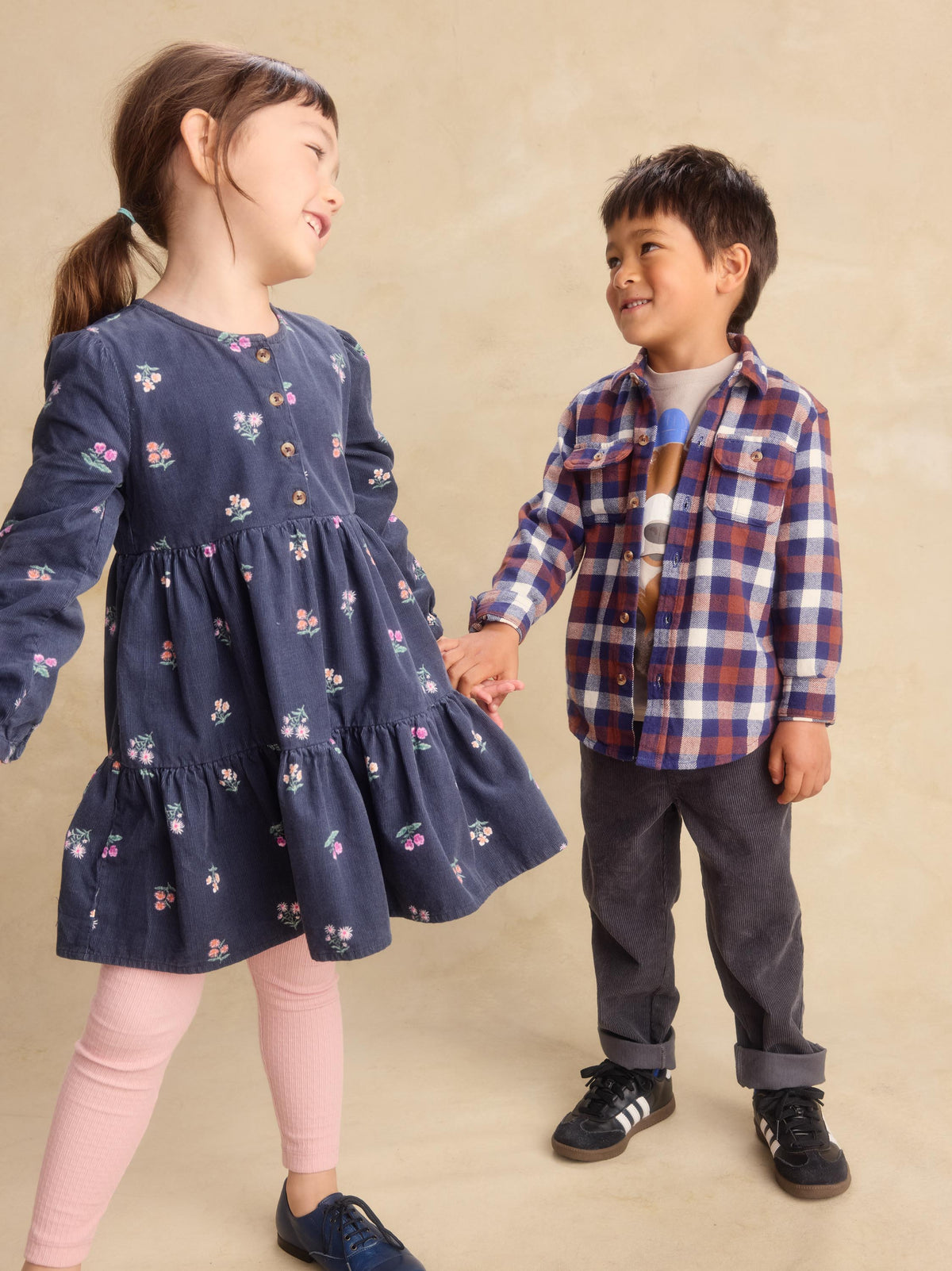 A young girl in a navy blue floral long-sleeve dress and a boy in a red, blue, and white plaid shirt are holding hands against a warm beige studio backdrop.