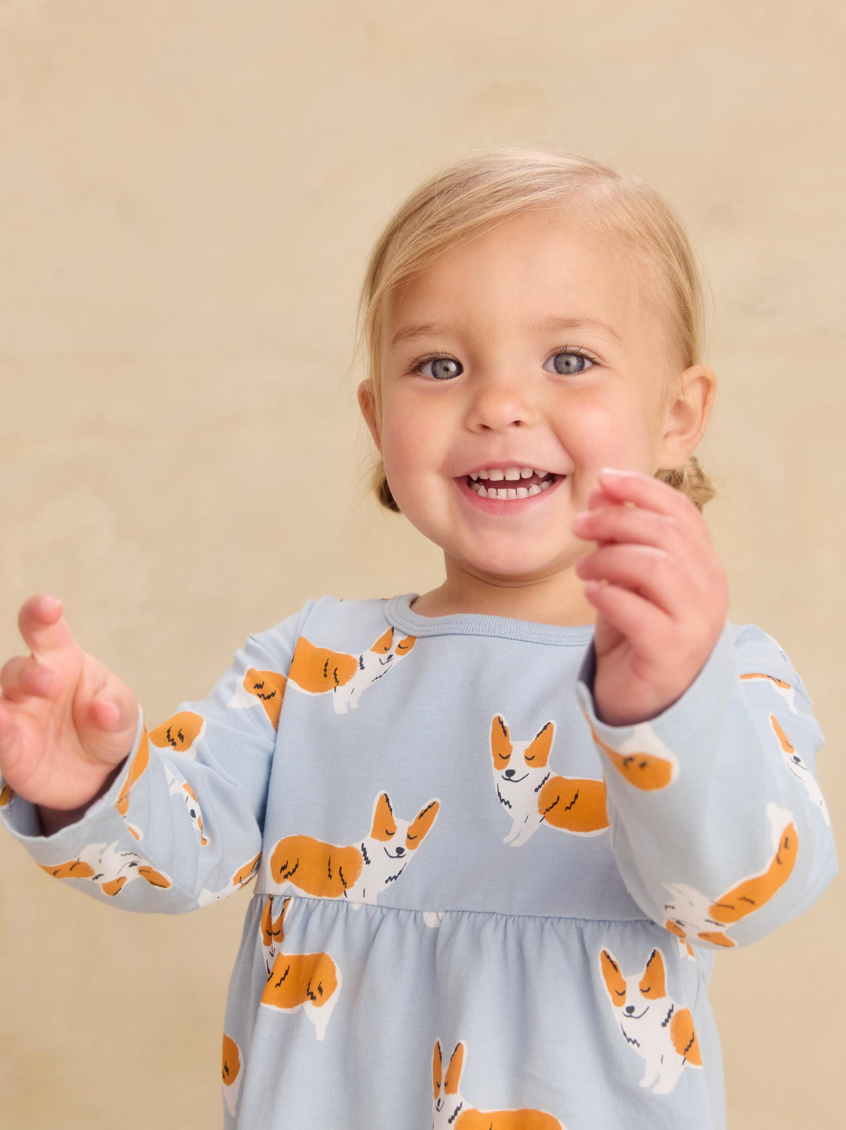 A smiling toddler girl wearing a light blue long-sleeve dress with an orange and white corgi dog print, posing against a warm beige background.
