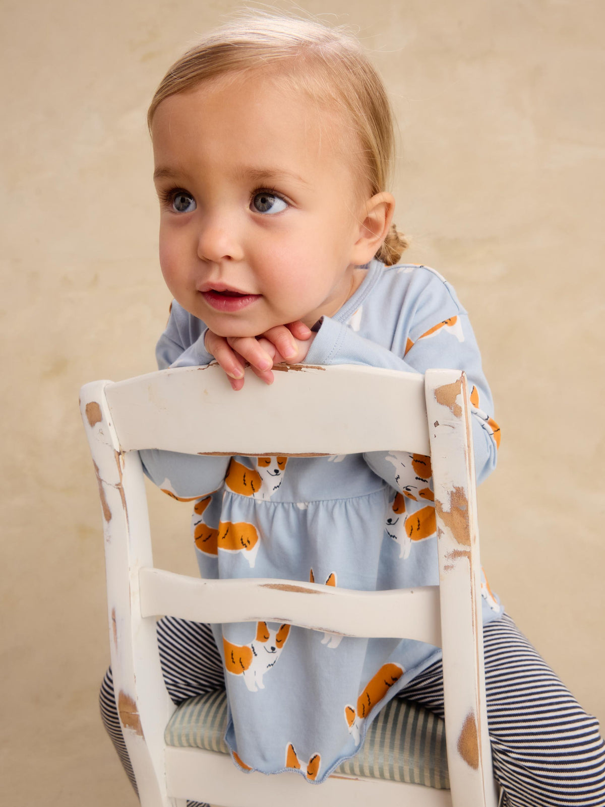 A toddler girl with blonde hair sits on a distressed white wooden chair, wearing a light blue long-sleeve dress with an orange and white dog print, paired with navy and white striped leggings, against a warm beige background.