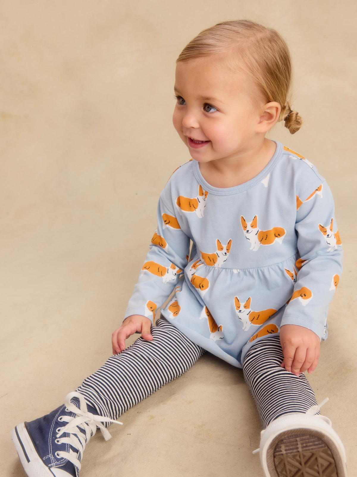 A toddler girl sitting on a beige floor, wearing a light blue long-sleeve top with an orange and white dog print, navy and white striped tights, and navy sneakers.