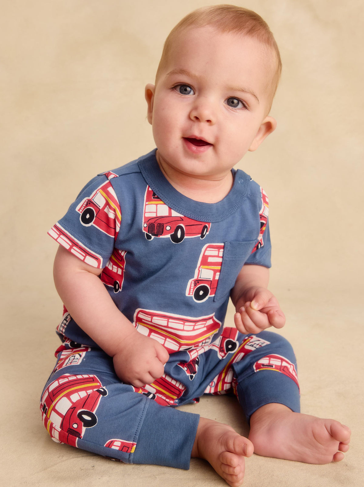 A baby sitting on a beige studio floor wearing a blue short-sleeve romper with a red and white bus print.