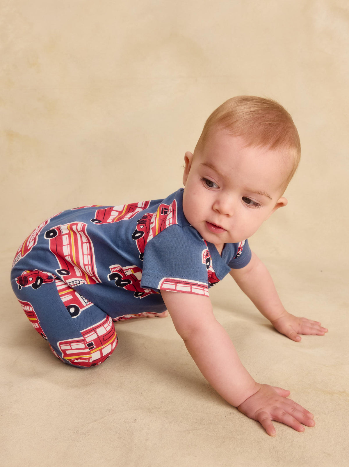 A baby crawling on a beige studio background, wearing a short-sleeve one-piece romper with a red and white bus pattern on a blue base.