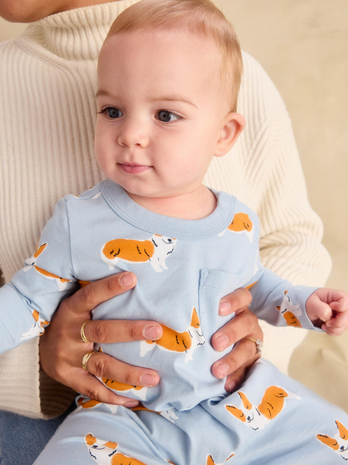 A baby wearing a light blue long-sleeve romper with an orange and white corgi print, held by an adult wearing a cream ribbed sweater.