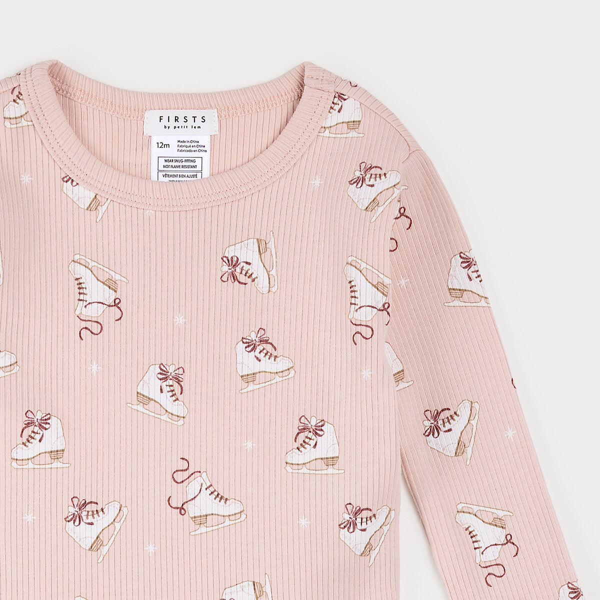 A close-up of a children's long-sleeve top in light pink waffle fabric, featuring an all-over print of white ice skates with brown laces and small white star accents. The brand label 'FIRSTS FOR KITTEN' is visible inside the collar.