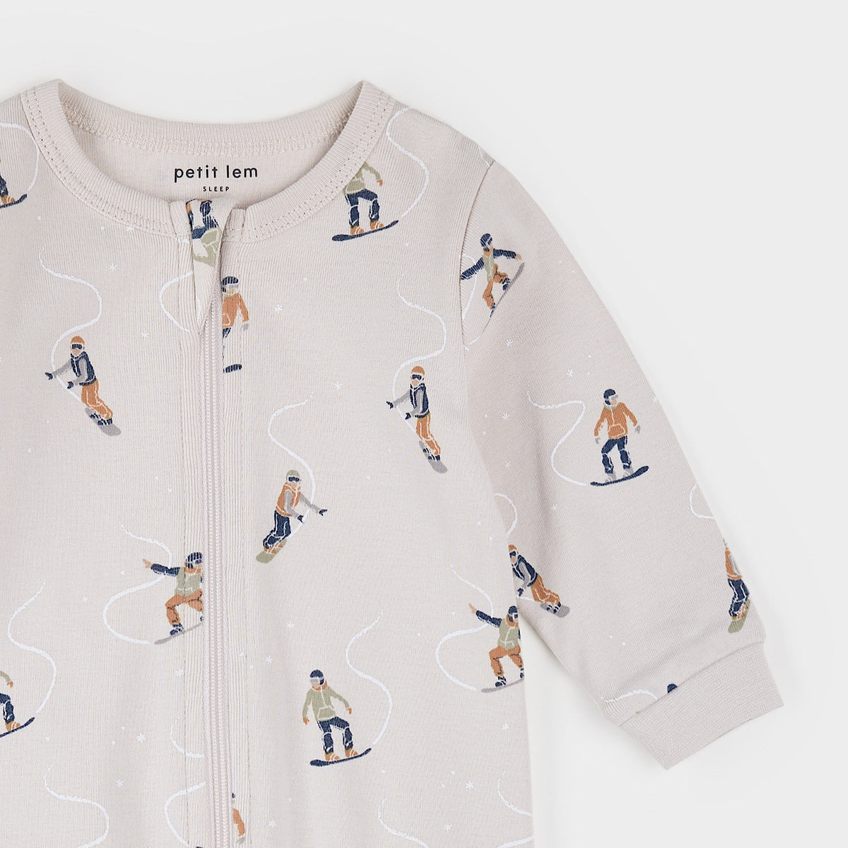 A close-up of a baby long-sleeve zip-up romper by Petit Lem Paris, featuring an all-over winter sports print of skiers and snowboarders on a light beige background.