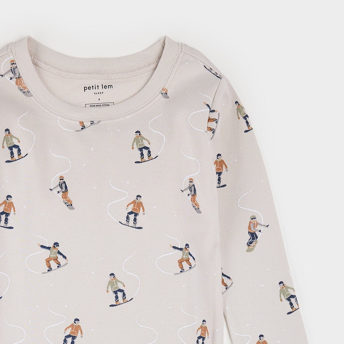 A close-up of a children's long-sleeve top in a light beige color, featuring an all-over print of cartoon snowboarders in orange, navy, and beige, with a visible 'petit lem' brand label inside the collar.