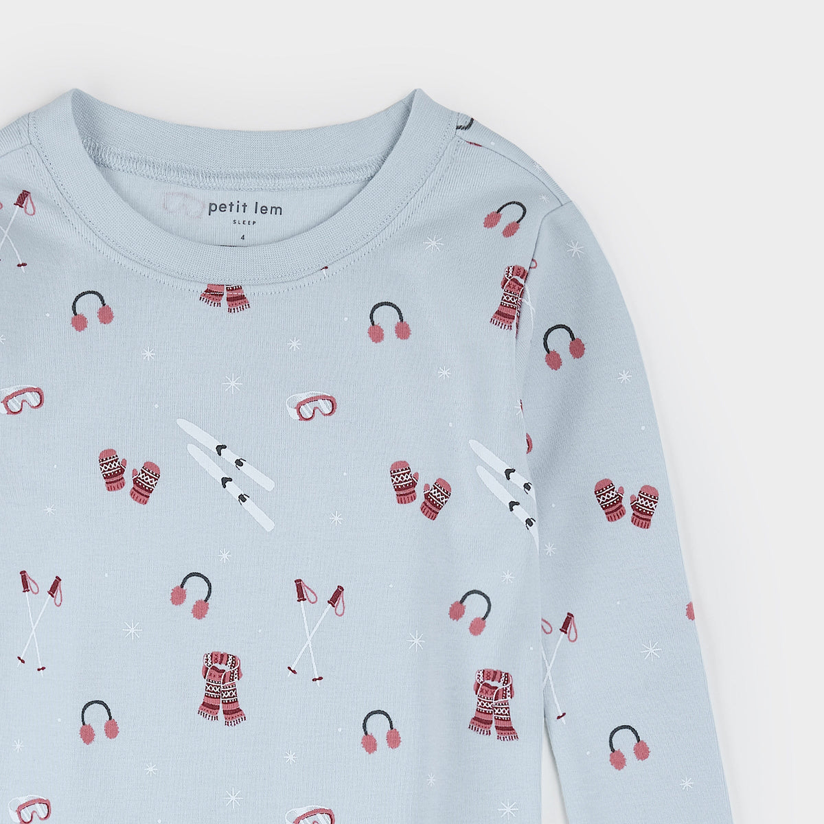 A light blue long-sleeve baby shirt with a winter sports print of red and white scarves, mittens, goggles, and skis, featuring the 'petit lem' brand label on the inside neck.