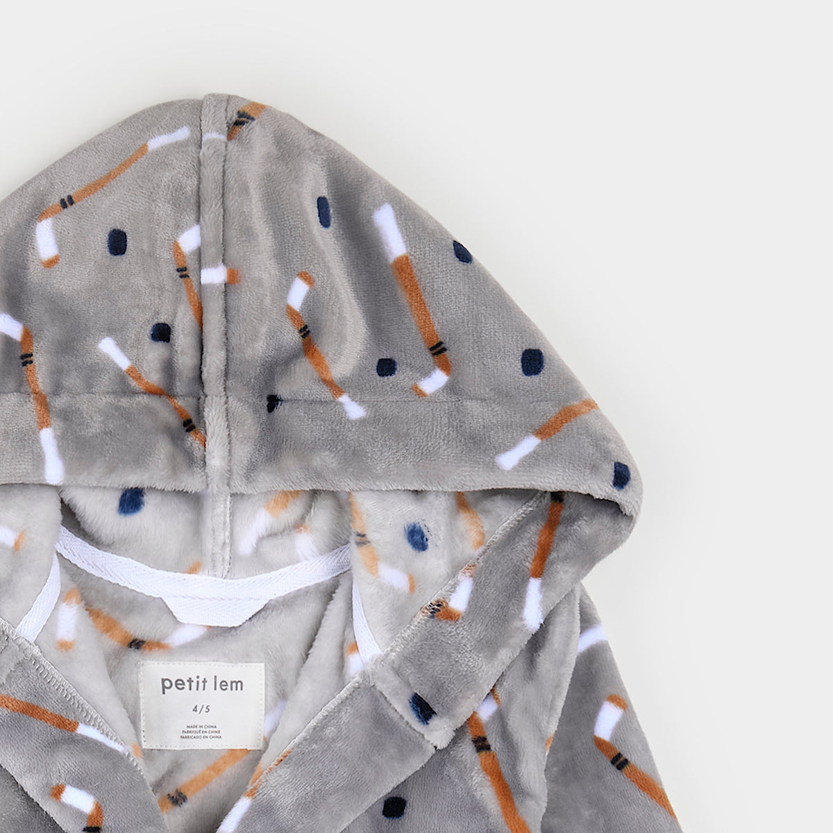 Close-up of a gray plush hooded robe with an allover hockey stick and puck print in orange, white, and navy, shown on a light neutral background. A brand label reading 'petit lem' is visible inside the collar.