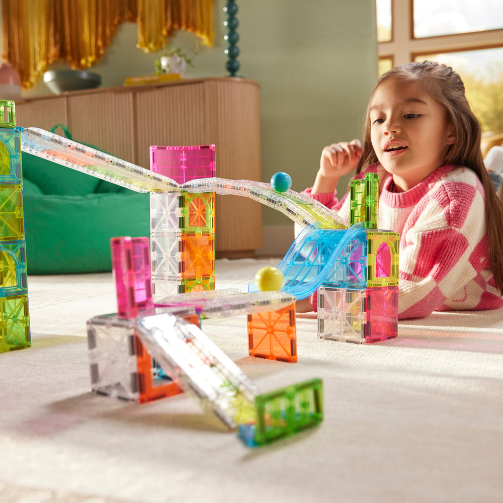 A young girl sits on a carpeted floor in a living room, playing with translucent magnetic building blocks.