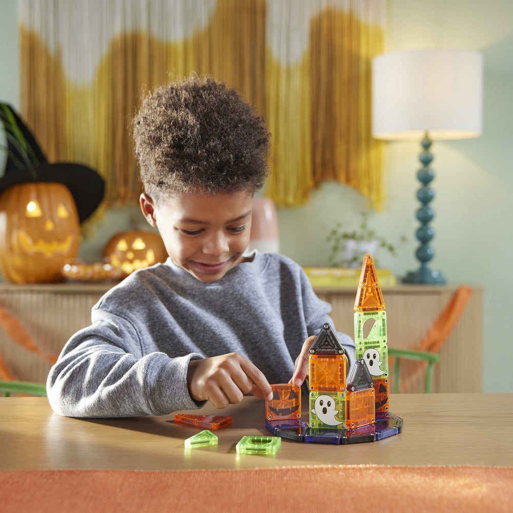 A young boy wearing a gray sweatshirt sits at a wooden table, playing with a colorful magnetic building toy castle made of translucent orange, green, and black pieces. The background shows a cozy, softly lit room with Halloween decorations, including a jack-o'-lantern and a witch hat on a shelf, and a blue-green spiral lamp on a table lamp stand.
