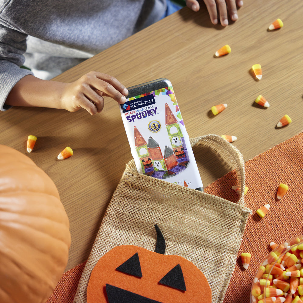 Person holding a smartphone with a Halloween-themed app over a burlap bag with a pumpkin design, surrounded by candy corn on a wooden table.