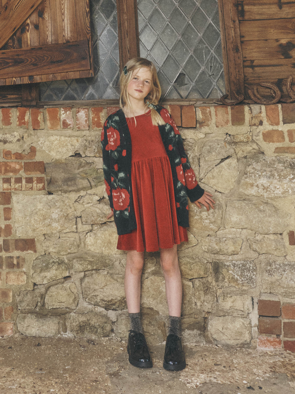 A young girl standing in front of a weathered stone wall, wearing a long dark knit cardigan with a red-and-green floral pattern over a reddish-orange dress, paired with dark boots and socks.