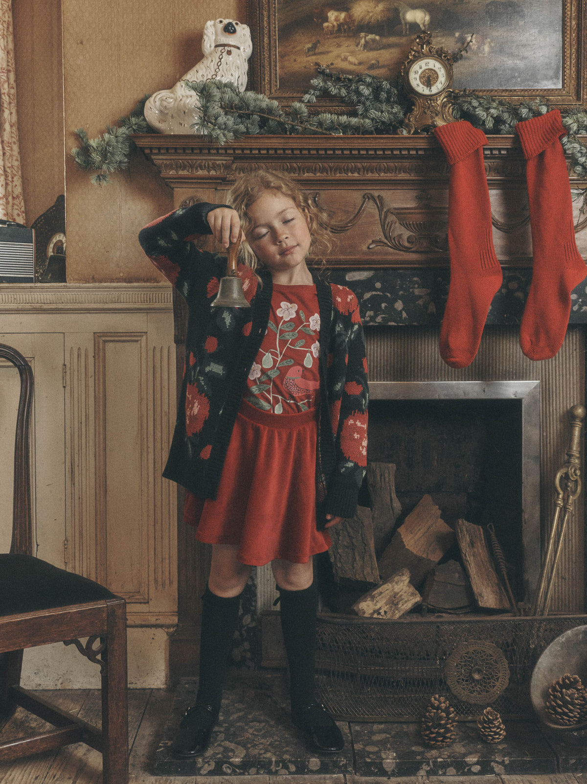 A young girl standing in a cozy, rustic living room next to a stone fireplace decorated with stockings and greenery, wearing a long dark knit cardigan with a red floral pattern over a reddish dress.