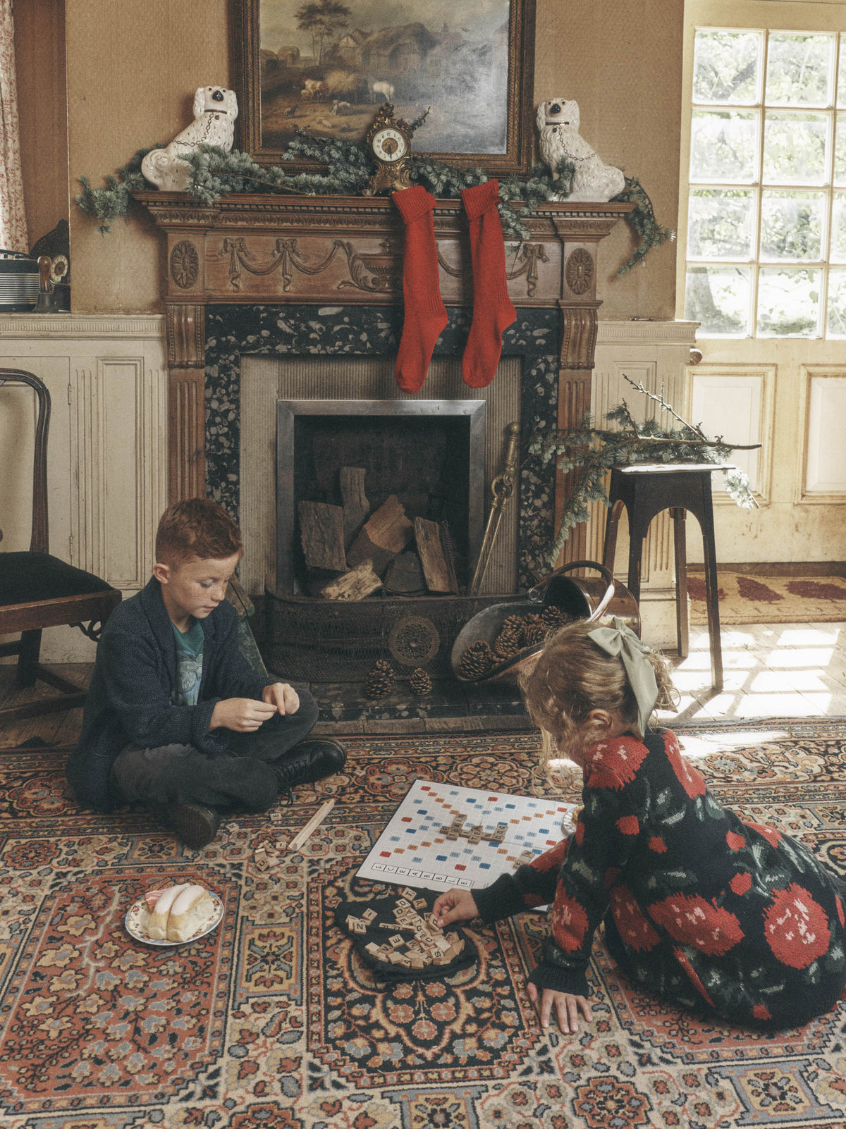 A young boy sitting on a patterned rug and a young girl looking at a book or activity in a cozy living room with a stone fireplace, stockings, and holiday greenery.