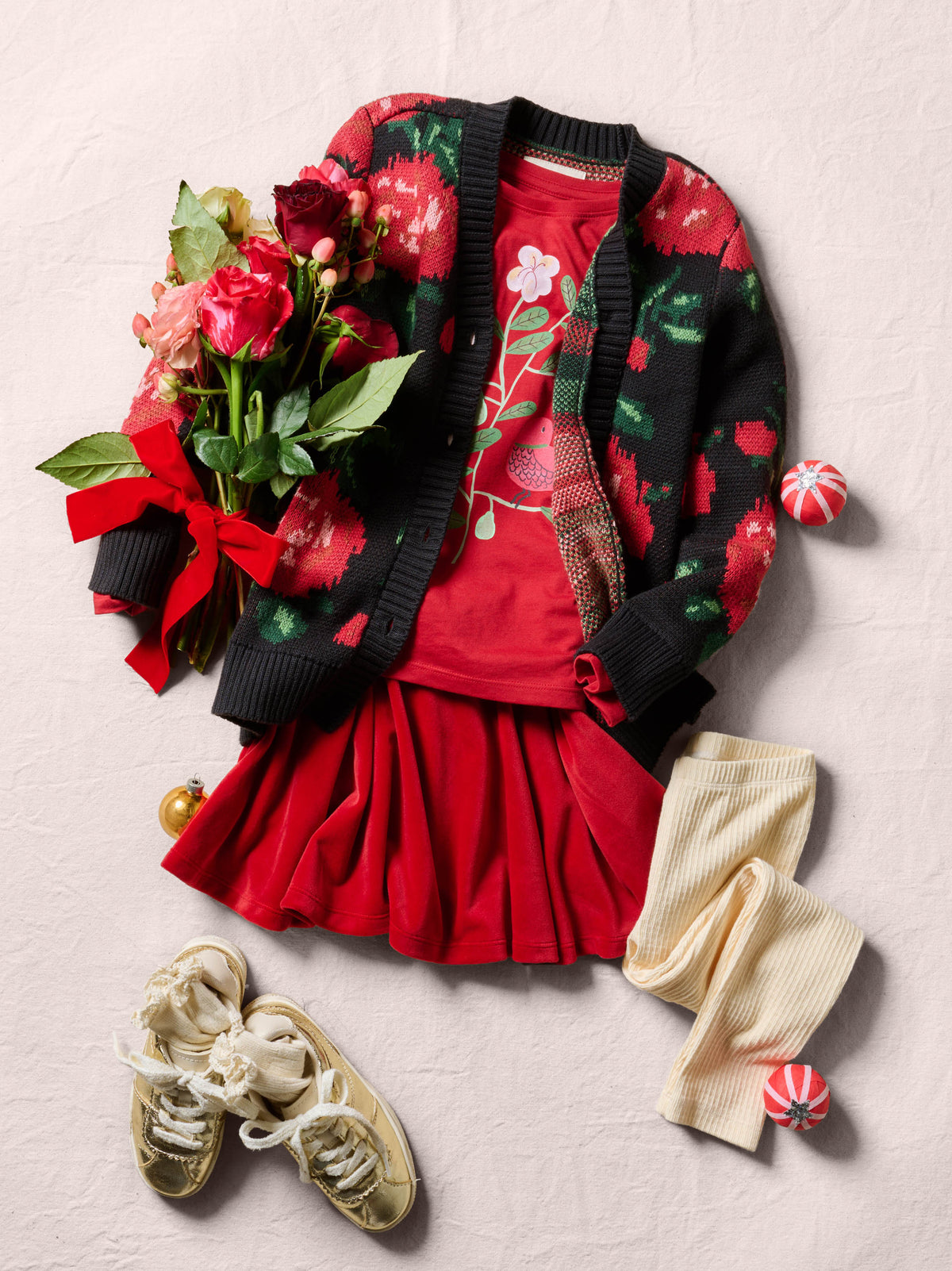 Flat lay of a dark knit cardigan with a red-and-green floral pattern, paired with a bright red skirt and top, a bouquet of red flowers with greenery, cream-colored tights, and gold shoes.