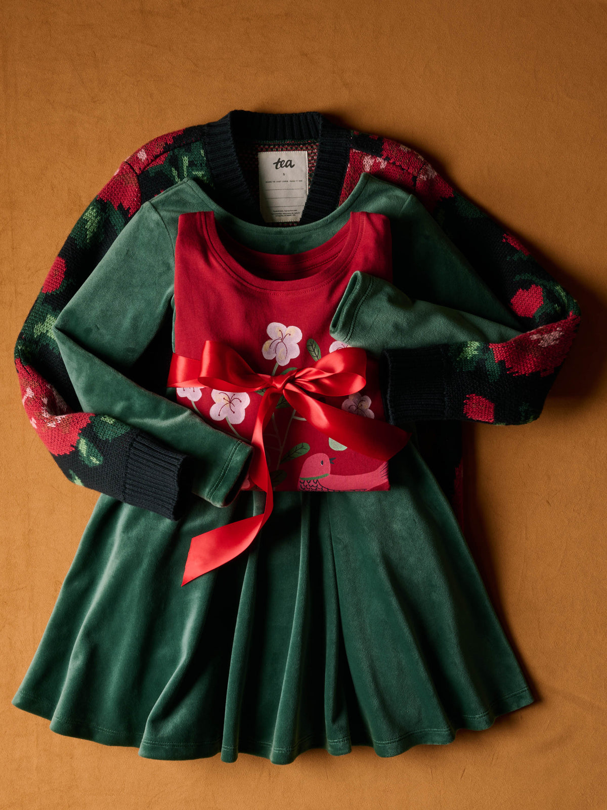 Flat lay of a dark knit cardigan with a red-and-green floral pattern, paired with a green dress featuring a red sash and floral accents, laid out on a warm reddish-brown textured background.