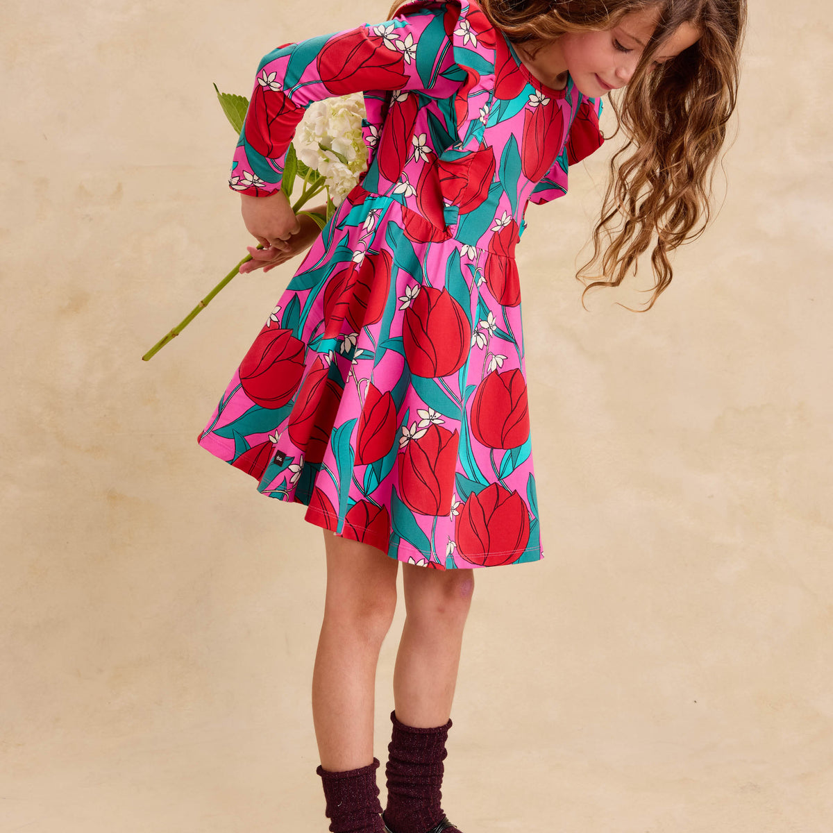 Ruffle Shoulder Ballet Dress - Tulip Twist in Pink by Tea Collection featuring long sleeves, a crew neckline, ruffled shoulder details, and an artist-drawn red and teal tulip floral pattern over a pink background, worn with dark socks and shoes while holding a flower.