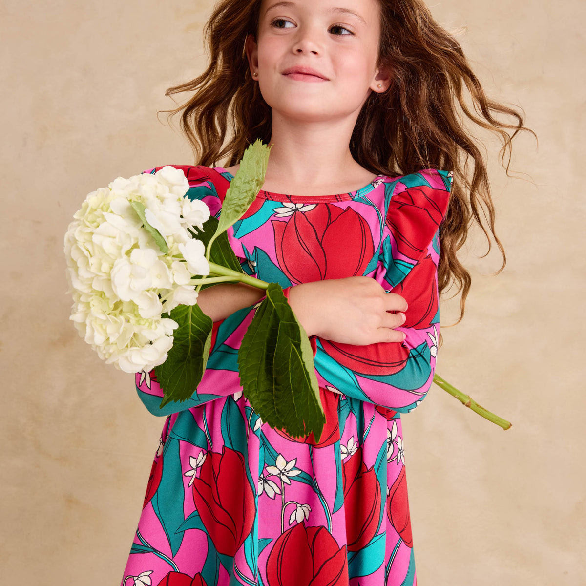 Ruffle Shoulder Ballet Dress - Tulip Twist in Pink by Tea Collection featuring long sleeves, a crew neckline, ruffled shoulder details, and an artist-drawn red and teal tulip floral pattern over a pink background, worn by a smiling child holding a large white flower.