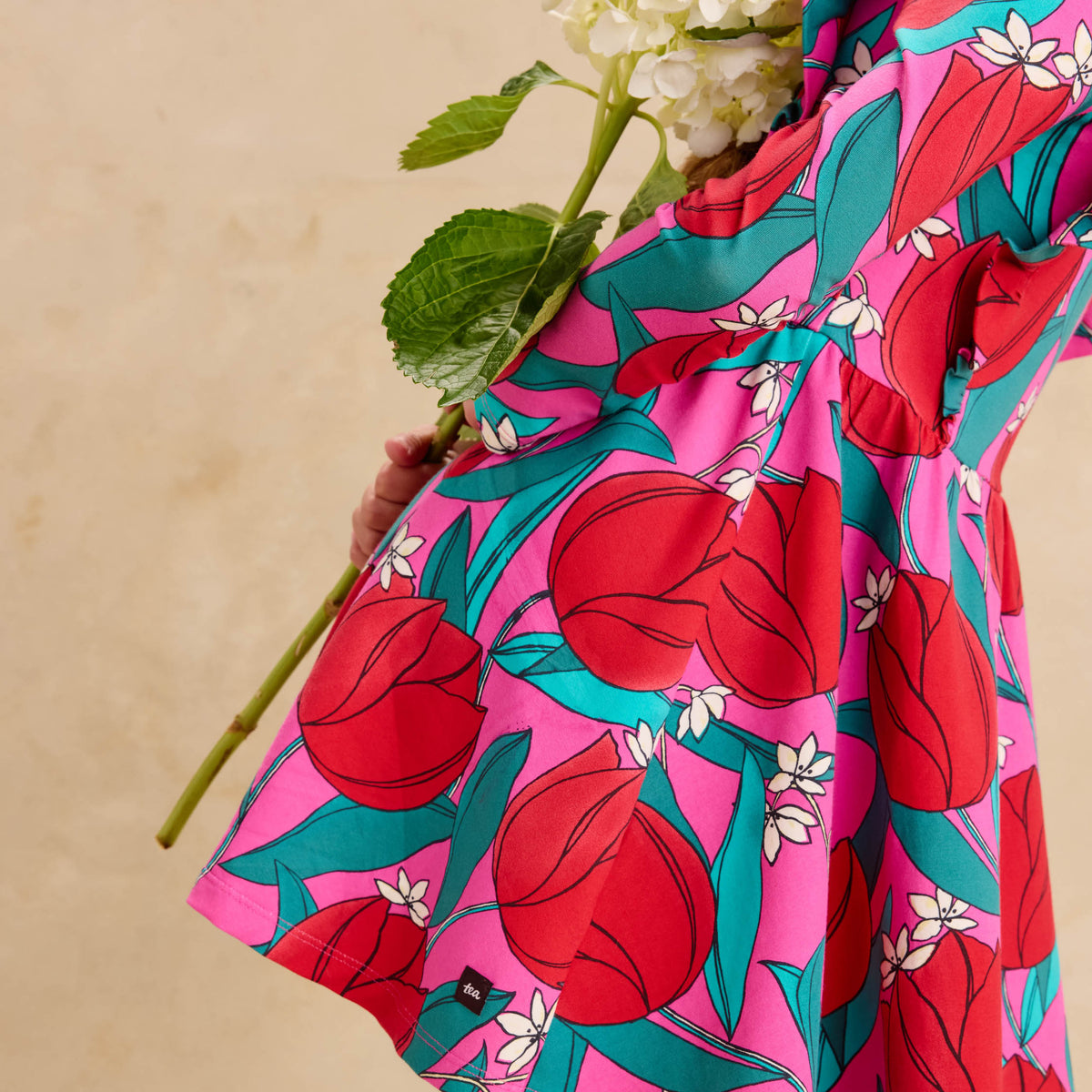 Ruffle Shoulder Ballet Dress - Tulip Twist in Pink by Tea Collection featuring long sleeves, a crew neckline, ruffled shoulder details, and an artist-drawn red and teal tulip floral pattern over a pink background, viewed from the side while holding a large white flower.