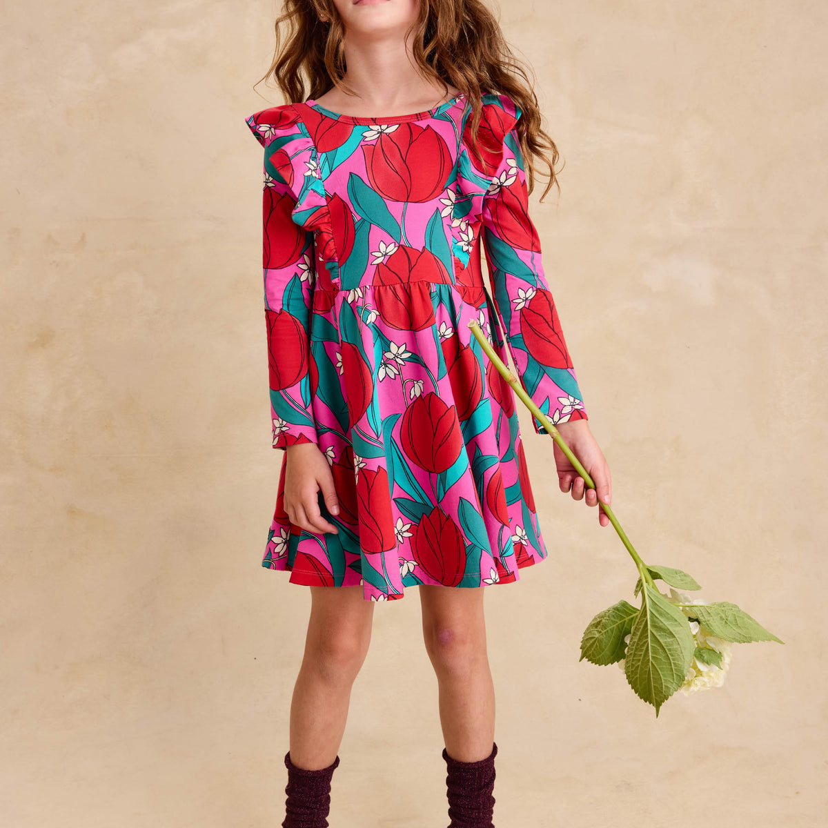 Ruffle Shoulder Ballet Dress - Tulip Twist in Pink by Tea Collection featuring long sleeves, a crew neckline, ruffled shoulder details, and an artist-drawn red and teal tulip floral pattern over a pink background, worn by a child standing while holding a large flower stem with green leaves.