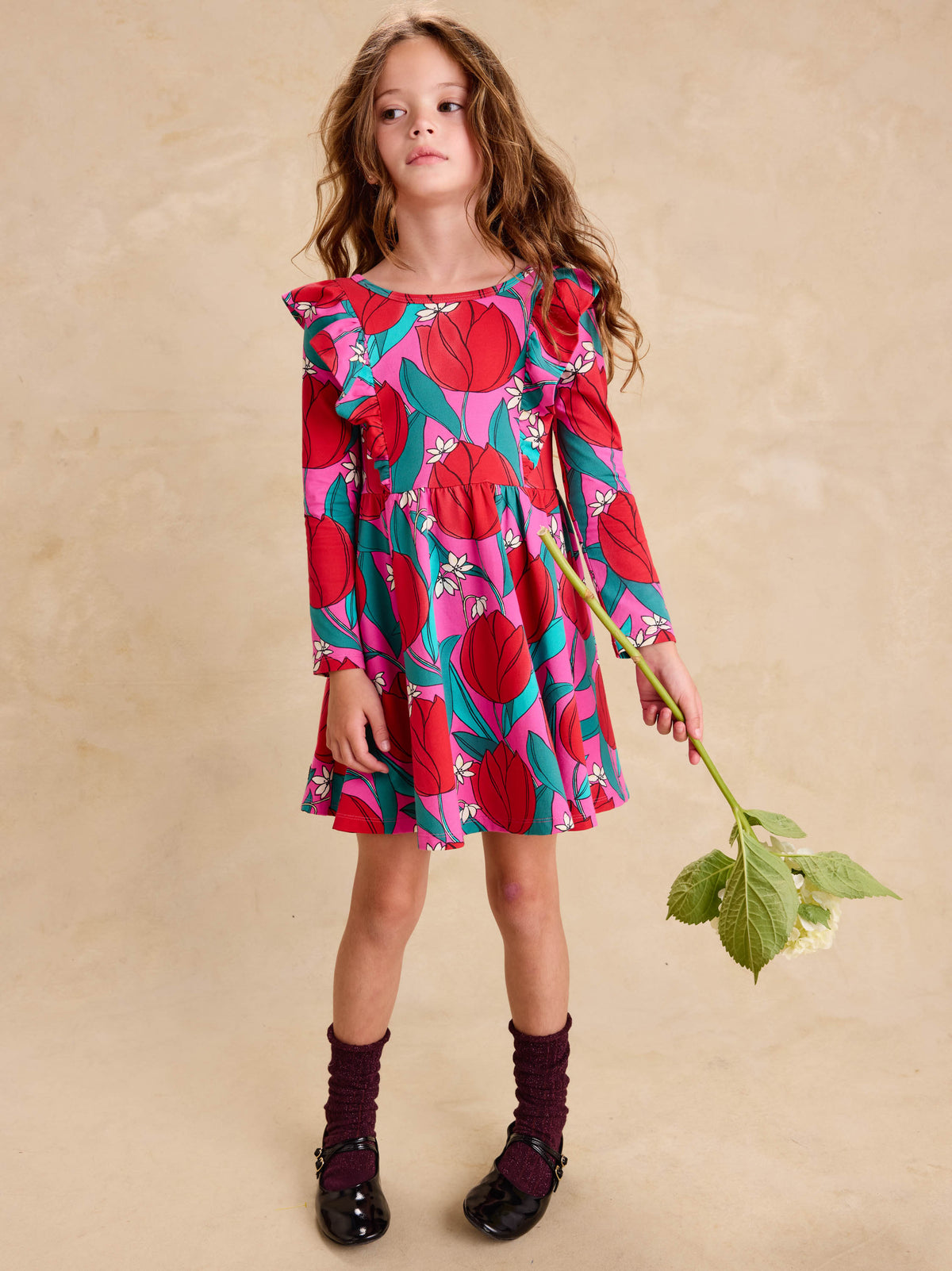 Ruffle Shoulder Ballet Dress - Tulip Twist in Pink by Tea Collection featuring long sleeves, a crew neckline, ruffled shoulder details, and an artist-drawn red and teal tulip floral pattern over a pink background, worn by a child standing while holding a large flower stem with green leaves.