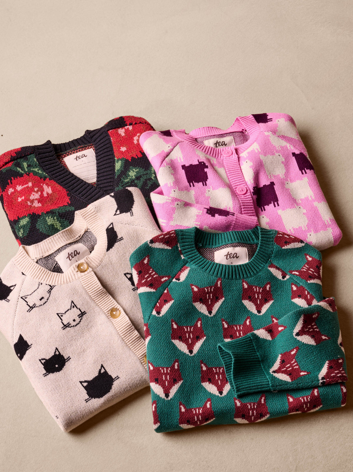 A flat lay view of multiple children's knitwear items folded and stacked together, including the emerald green Family Fox Sweater - Foxwood Fair Isle by Tea Collection, alongside a beige sweater, a pink patterned cardigan, and a dark sweater, on a textured surface.