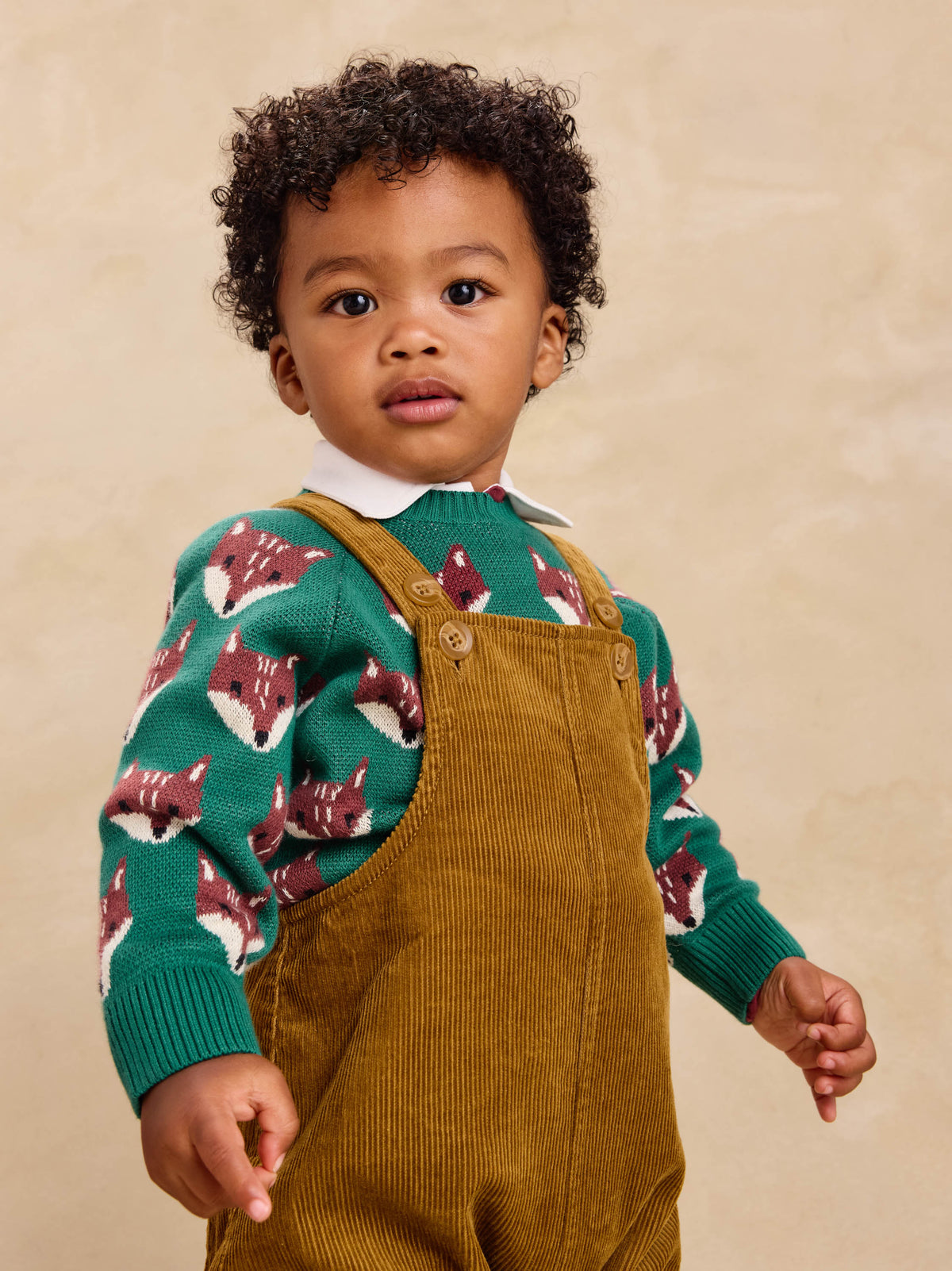 A front view of the Family Fox Sweater - Foxwood Fair Isle by Tea Collection, featuring an emerald green knit background patterned with rows of repeating, stylized red-and-white fox faces across the chest and sleeves, set against a plain white background.